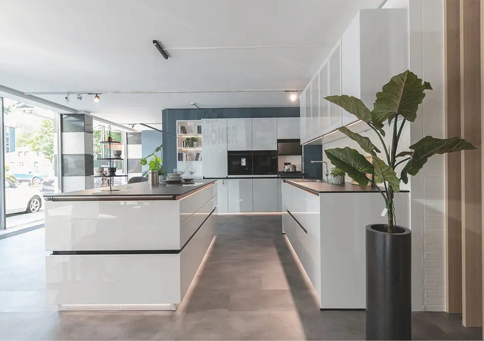 Helle Küche mit großer Kochinsel, weißen Fronten und Arbeitsfläche, rechts große Pflanze im Topf im Showroom